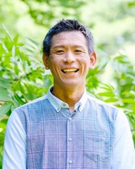 木々の緑を背景に笑顔で立つ潮田英之